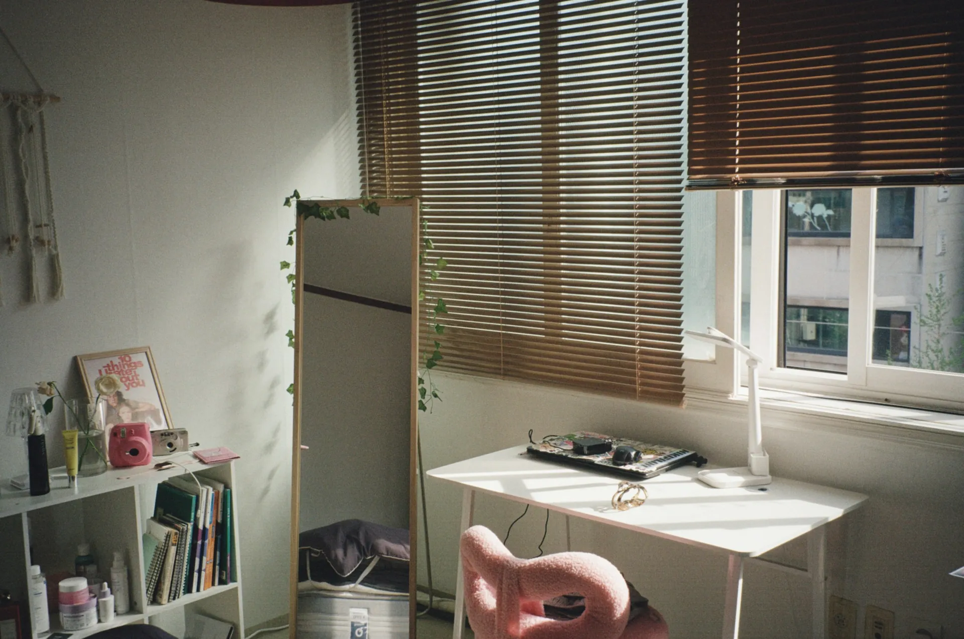 Child's study desk with a pink chair beneath window blinds and a low bookshelf