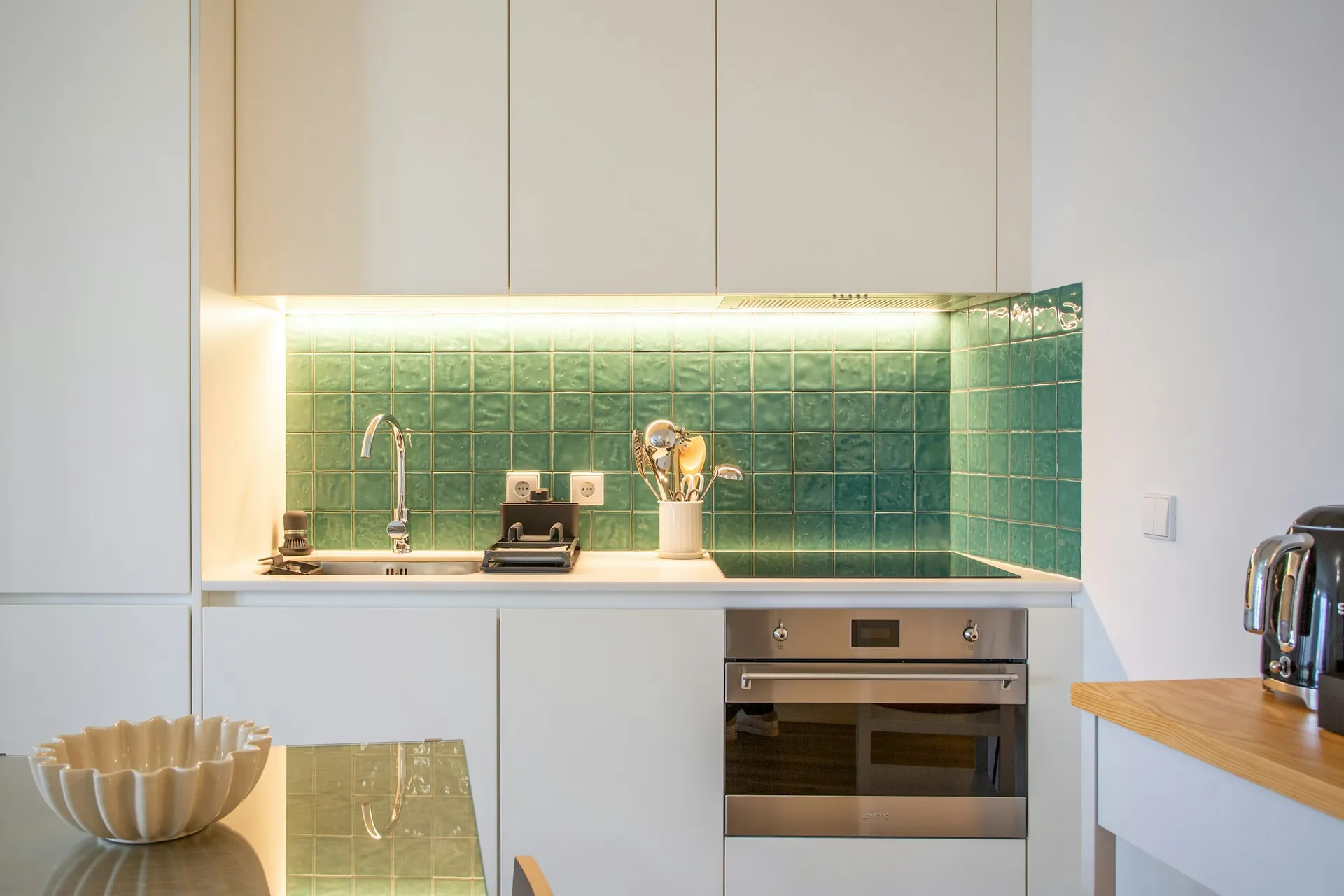 Green tiled kitchen backsplash lit by under-cabinet lights above a white counter